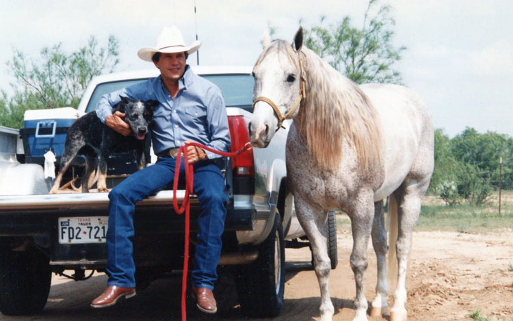 George Strait sits on the tailgate of the pickup petting a blue heeler dog and holding the lead rope of a grey horse standing beside the truck.